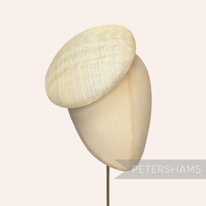 May include: A cream-colored, woven hat with a rounded crown, displayed on a mannequin head. The hat has a textured appearance and is likely made of natural fibers. The mannequin head is a neutral beige color. The word "PETERSHAMS" is visible in the lower right corner.
