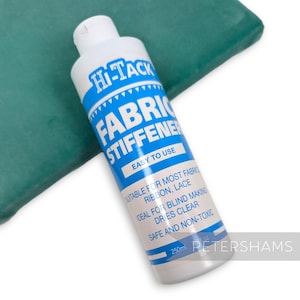 May include: A blue bottle of Hi-Tack Fabric Stiffener, a clear liquid for stiffening fabric. The bottle is labelled with instructions for use, including "Easy to Use", "Suitable for most fabrics, ribbon, lace", "Ideal for blind making", "Dries clear", "Safe and non-toxic", and "250ml". The bottle is sitting on a green surface.