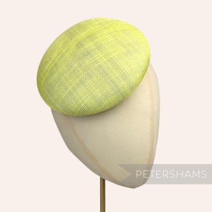 May include: A bright yellow, round hat made of woven straw, displayed on a white hat stand. The hat is tilted slightly to the side. The stand has a gold metal rod.