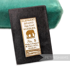 May include: A black rectangular package of "ELEPHANT BRAND Egg Eyed NEEDLES" with a gold label. The label features an elephant illustration and reads "No. 5 Made in England W. HALL & Co., STUDLEY. HARNESS". A teal green velvet surface is in the background.