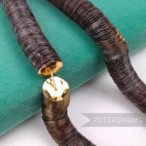 May include: A close-up of a brown sequin necklace with gold-colored clasps. The necklace is made of small, round sequins strung together. The clasp is a round, gold-colored metal piece. The necklace is displayed on a teal-colored surface. The text "PETERSHAMS" is visible.