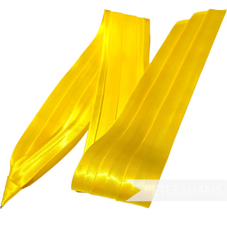 Pode incluir: Uma fita de cetim amarelo brilhante, dobrada e disposta de forma decorativa. A fita &eacute; lisa e brilhante, com um leve brilho. &Eacute; uma fita vers&aacute;til que pode ser usada para uma variedade de prop&oacute;sitos, como embrulhar presentes, decorar artesanato ou adicionar um toque de eleg&acirc;ncia a qualquer projeto.