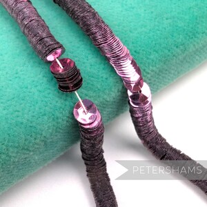 May include: Close-up of two strands of dark purple sequins on a teal background. The sequins are circular and sewn together with white thread. The sequins are a mix of matte and shiny finishes.