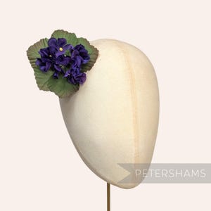Può includere: Un manichino per cappello color crema con un gruppo di fiori viola e foglie verdi. I fiori hanno centri gialli. La parola "PETERSHAMS" è stampata in bianco in basso a destra.