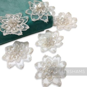May include: Close-up of several white, beaded floral appliques. The appliques are made of delicate lace and feature intricate beadwork in the centre. They are arranged on a teal green velvet surface. The appliques are likely for embellishing clothing or accessories.