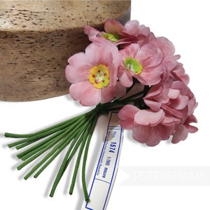 May include: A bunch of 10 pink silk flowers with green stems. The flowers have a yellow center and are arranged in a bouquet. The flowers are labeled with a tag that reads "No. 1574 1/7655 mauve Made in German Democratic Republic."