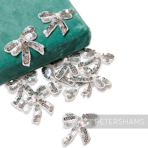 May include: Silver sequin bow embellishments scattered on a white surface and a teal green velvet surface. The bows are made of silver sequins and white thread. The image includes the text "PETERSHAMS".
