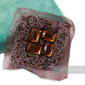 May include: A square, beaded embellishment with four amber-colored, rectangular gems at the center. The beads are a mix of brown, gold, and purple. The embellishment is on a sheer, mauve fabric, with a teal fabric in the background.