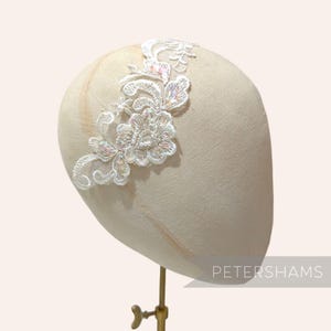 May include: A white lace headband with floral and leaf designs, adorned with iridescent sequins. The headband is displayed on a mannequin head. The word "PETERSHAMS" is printed in white on the lower right.