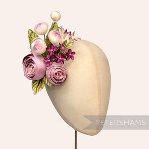 Op de afbeelding: Een bloemenhoofddeksel met roze stoffen rozen, kleine paarse bloemen en groene bladeren. De opstelling is bevestigd op een mannequinhoofd. Het woord "PETERSHAMS" staat in het wit afgedrukt aan de rechterkant van de afbeelding.