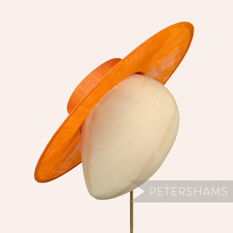 May include: A large, bright orange, wide-brimmed hat made of woven straw. The hat is sitting on a white hat form.