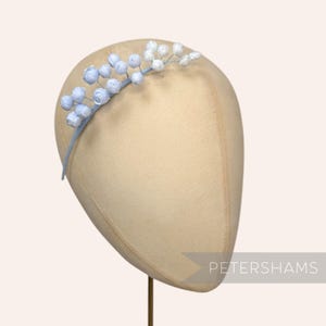 May include: A light blue headband with floral accents. The headband features a delicate design of small, light blue and white flowers and berries. The headband is displayed on a mannequin head.