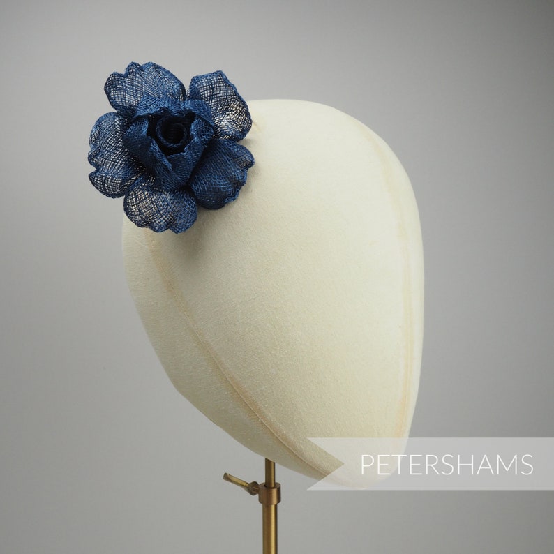 May include: A navy blue fabric flower, resembling a rose, is attached to a cream-colored hat form. The flower is made of a textured material. The hat form is mounted on a gold-colored stand. The text "PETERSHAMS" is visible.