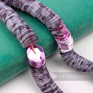 May include: A close-up of a necklace made of stacked, round, metallic sequins in shades of purple and silver. The necklace is resting on a teal green velvet surface. The sequins are connected by a thin, pink thread. The word "PETERSHAMS" is visible in the bottom right corner.