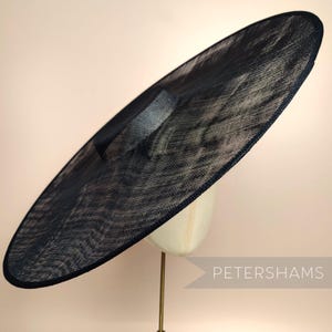 May include: A large, black, wide-brimmed hat made of a fine, woven material. The hat is sitting on a stand and is angled slightly to the side.
