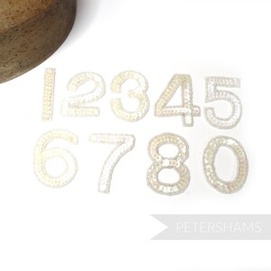 May include: Ten white pearl-like sequined numbers, 1 through 0, on a white background. The numbers are approximately 5 cm tall.