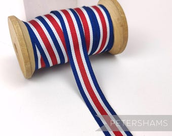 May include: A wooden spool of striped ribbon in red, white, and blue. The ribbon is unwound, showing the pattern of vertical stripes. The spool is made of light-colored wood, and the ribbon is likely used for crafts or decoration.