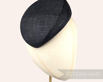 May include: A black, round, woven hat sits on a white hat stand. The hat is angled slightly to the side. The stand is on a gold metal base. The text "PETERSHAMS" is visible in the lower right corner of the image.