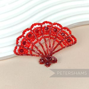 Può includere: Un ventaglio a forma di conchiglia di mare in perle rosse, realizzato con piccole perle rosse e filo metallico. Il ventaglio è aperto e presenta un design delicato e intricato.