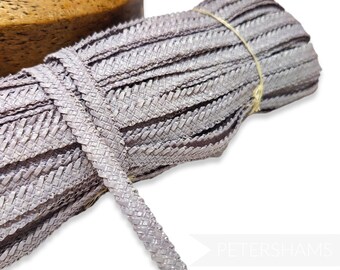 May include: A roll of light grey braided trim, woven from a flat, textured material. The trim is approximately 1.2 cm wide and is perfect for adding a touch of elegance to any project.
