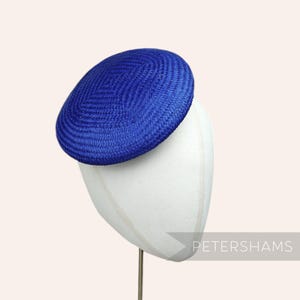 May include: A bright blue, round, flat hat made of woven straw. The hat is displayed on a white hat form.