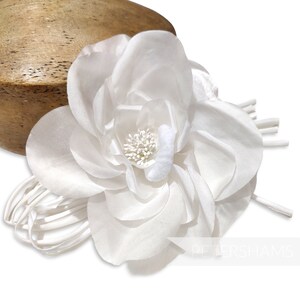 May include: A large, white fabric flower with multiple layers of petals and a textured center. The flower is attached to a bundle of thin, white stems. The background is white, with a wooden object partially visible.