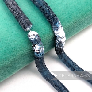 May include: Close-up of two strands of blue and silver sequins. The sequins are circular and sewn together to form a flexible strand. The sequins transition from dark blue to silver. The word "PETERSHAMS" is in the bottom right corner.