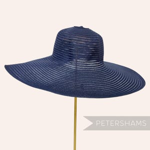 May include: A navy blue sun hat with a wide brim and a structured crown. The hat has a striped pattern and is displayed on a gold stand. The text "PETERSHAMS" is visible on the right side of the image.