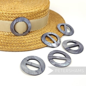 以下が含まれることがあります： 中央にバーが付いたグレーの真珠貝製の帽子留めが6つ。留め具は、ベージュのリボンが付いた麦わら帽子を囲むように半円形に配置されています。