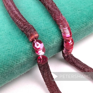 May include: Close-up of a burgundy sequin trim with a matching sequin clasp. The trim is made of small, round sequins sewn onto a fabric backing. The background is a teal green fabric.