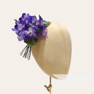 Könnte beinhalten: Ein lilafarbener Blumenhaarschmuck mit grünen Blättern auf einem beigefarbenen Mannequin-Kopf. Das Mannequin steht auf einem goldenen Ständer.