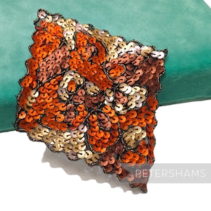 May include: A close-up of a square-shaped sequined applique with a swirling pattern in shades of orange, brown, and gold. The applique is on a green background.