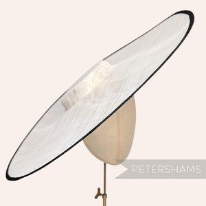 Peut inclure: Un chapeau à large bord en matériau tissé blanc semi-transparent avec un fin liseré noir sur le pourtour. Le chapeau présente une calotte structurée et est présenté sur un mannequin de couleur neutre. Le texte PETERSHAMS est visible.