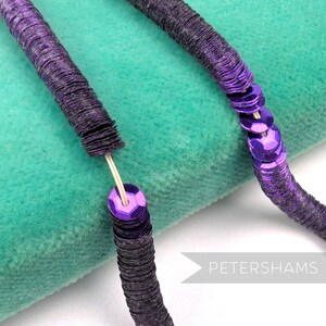 May include: Close-up of a purple sequin trim. The trim is made of small, round, metallic sequins sewn together in a line. The sequins are a vibrant purple color and have a shiny finish. The trim is on a teal green velvet surface.