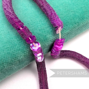 May include: Close-up of a purple sequin necklace with a matching cord. The necklace is made of small, round, iridescent sequins and is displayed on a teal green surface. The word "PETERSHAMS" is visible in the lower right corner.