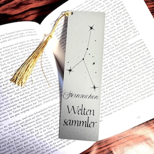 Könnte beinhalten: Ein Lesezeichen mit einem Sternbild-Design und den deutschen Worten "Sternzeichen Weltensammler" (Sternzeichen-Weltsammler). Es hat eine goldene Quaste und liegt auf einem Buch.