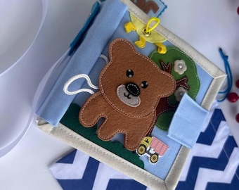 Puede incluir: Libro de actividades de fieltro hecho a mano con un diseño de oso pardo, árbol y camión. El libro presenta colores azul, verde y beige, con un cierre de cuerda blanca. Una etiqueta de fieltro azul con el nombre "John" es visible.