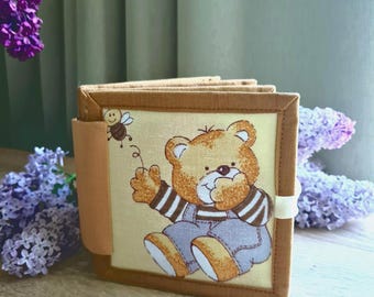 May include: A fabric book with a cartoon bear illustration. The bear is wearing overalls and a striped shirt. The book has a light brown border and a beige background. A small bee is also pictured. The book is closed with a white elastic band.