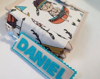 Puede incluir: Un libro cubierto de tela con un diseño de dibujos animados de nativos americanos. El marcapáginas dice "DANIEL" en letras azules. El libro tiene un borde beige y un fondo blanco con flechas y tipis.