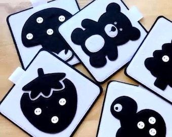 May include: Five black and white felt squares with different shapes and button embellishments. The shapes include a mushroom, a bear, a strawberry, a turtle, and a tree.