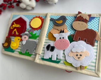 May include: A felt book for children featuring farm animals. The book has a red barn, a yellow sun, a gray goat, a yellow duck, a brown horse, a white cow, a white sheep, and a blue bird.