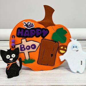 May include: An orange felt pumpkin with the words "HAPPY BOO" and a door, a black cat, and a white ghost. The pumpkin has a brown stem and green leaf. The cat has orange accents. The ghost has blue eyes.