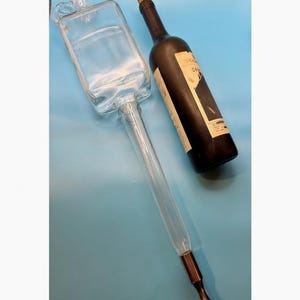 May include: A clear glass wine aerator and a dark wine bottle with a cork stopper. The aerator has a rectangular chamber and a long, slender tube with a metal tip. The wine bottle has a label.