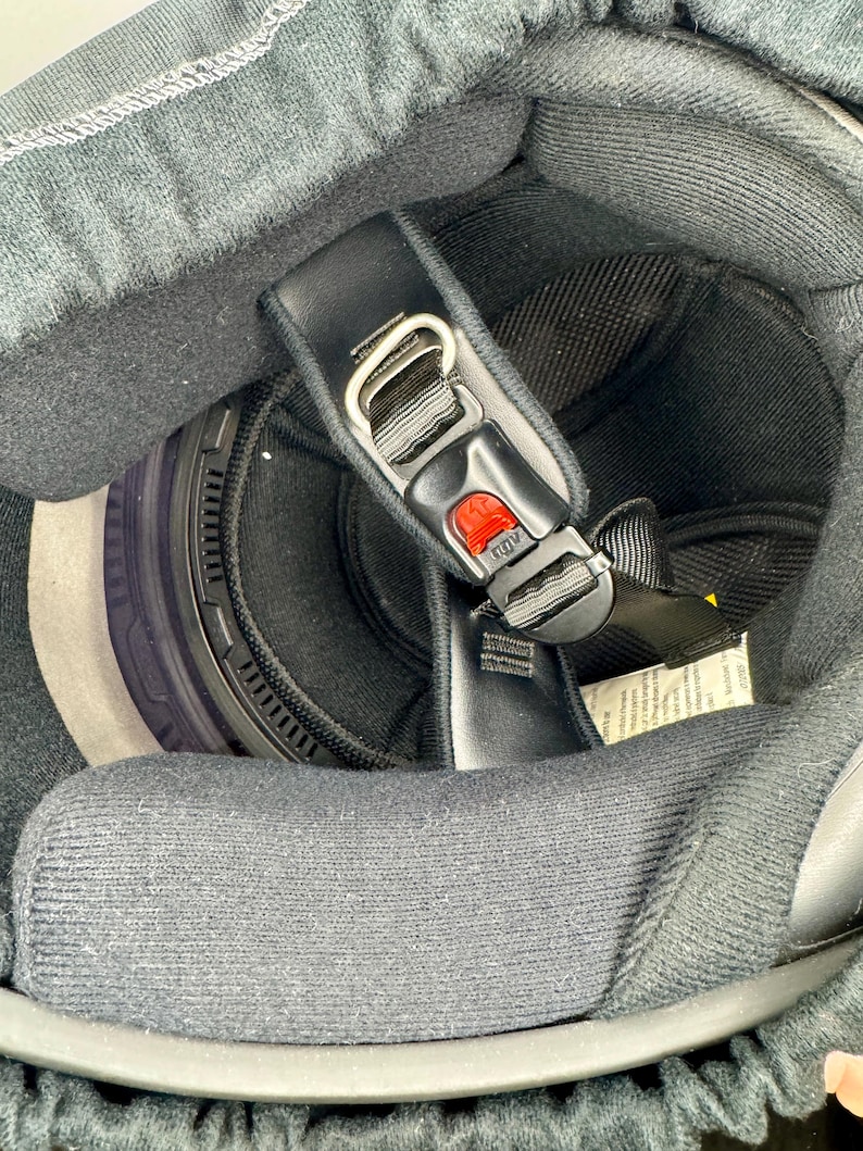 Peut inclure: Vue int&eacute;rieure d'un casque de moto noir. Le casque est dot&eacute; d'une jugulaire noire avec un bouton de d&eacute;verrouillage rouge, d'une doublure int&eacute;rieure noire et d'un tour de cou noir. Le rembourrage int&eacute;rieur du casque est gris.