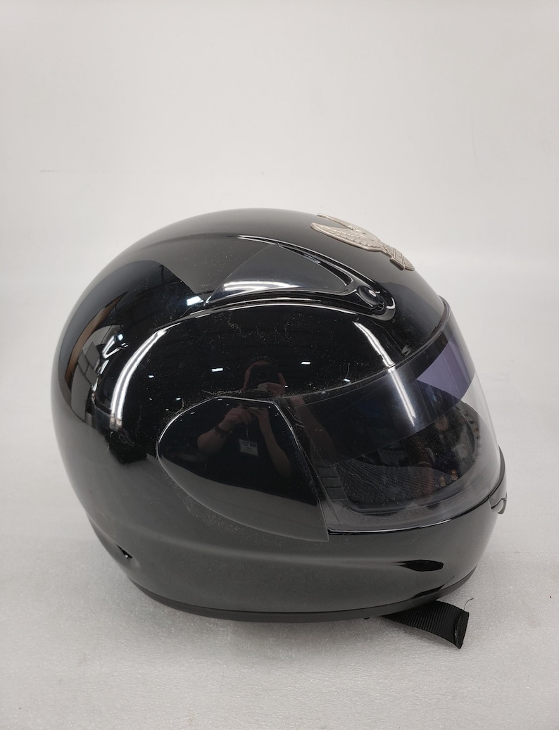 Peut inclure: Casque de moto noir brillant avec une visi&egrave;re teint&eacute;e. Le casque a un design &eacute;pur&eacute; et une jugulaire. Un embl&egrave;me argent&eacute; est visible sur le dessus du casque.