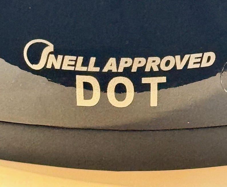 Peut inclure: Gros plan sur une surface bleu fonc&eacute; avec le texte blanc "Snell Approved DOT". Le texte est en police sans-serif. L'image est bien &eacute;clair&eacute;e et le texte est clair et facile &agrave; lire.