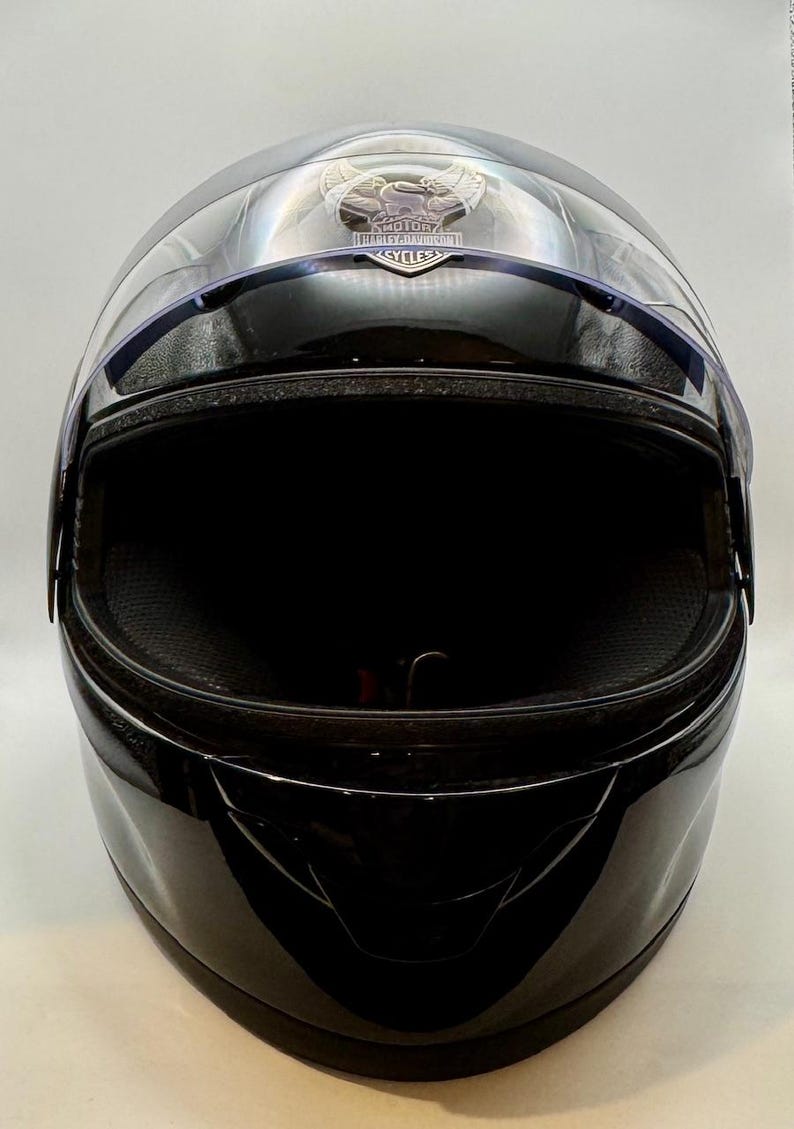 Peut inclure: Casque de moto noir brillant avec une visi&egrave;re transparente. Le casque pr&eacute;sente un logo Harley-Davidson sur le dessus. La visi&egrave;re est l&eacute;g&egrave;rement teint&eacute;e et se courbe &agrave; l'avant. L'int&eacute;rieur est noir.