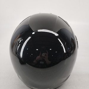 Peut inclure: Un casque de moto noir brillant. Le casque a une forme arrondie avec une surface lisse et r&eacute;fl&eacute;chissante. L'image montre l'arri&egrave;re du casque, avec une l&eacute;g&egrave;re courbe en haut et un &eacute;l&eacute;ment de design subtil pr&egrave;s de la base.