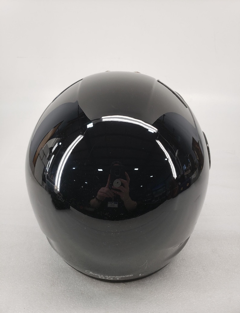 Peut inclure: Un casque de moto noir brillant. Le casque a une forme arrondie avec une surface lisse et r&eacute;fl&eacute;chissante. L'image montre l'arri&egrave;re du casque, avec une l&eacute;g&egrave;re courbe en haut et un &eacute;l&eacute;ment de design subtil pr&egrave;s de la base.