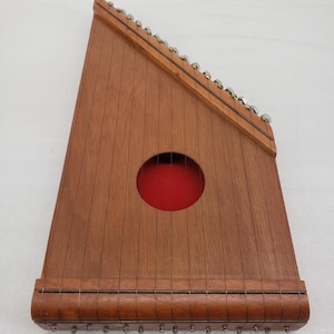 May include: A wooden autoharp with a triangular shape. The instrument has a dark brown finish, a round sound hole with a red interior, and multiple strings. Metal tuning pegs are visible along the top edge.